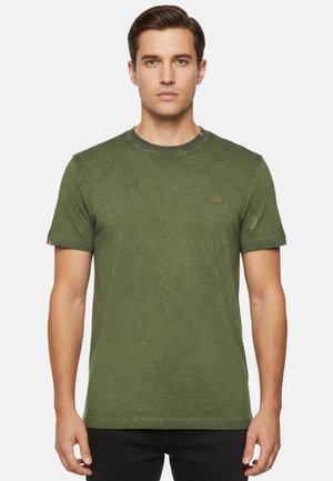 Giovane uomo in piedi di fronte, indossa una t-shirt verde oliva chiaro a maniche corte e pantaloni neri su uno sfondo neutro.