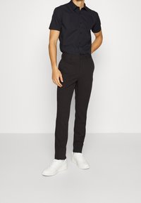 Schwarzes kurzärmeliges Hemd mit Knöpfen, kombiniert mit schmal geschnittenen schwarzen Hosen und weißen Sneakern, mit einer glatten Textur und minimalistischem Design.