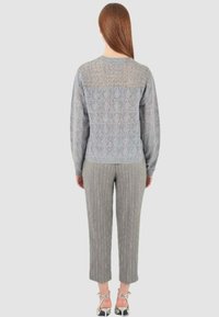 Donna con capelli lunghi che indossa un maglione grigio a maglia, pantaloni a cavallo corto grigio con righe e sandali argentati con tacco, di spalle su uno sfondo neutro.