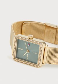 Montre carrée dorée avec un bracelet en maille, cadran vert, accents en cristal autour de la lunette et une petite couronne pour le réglage.
