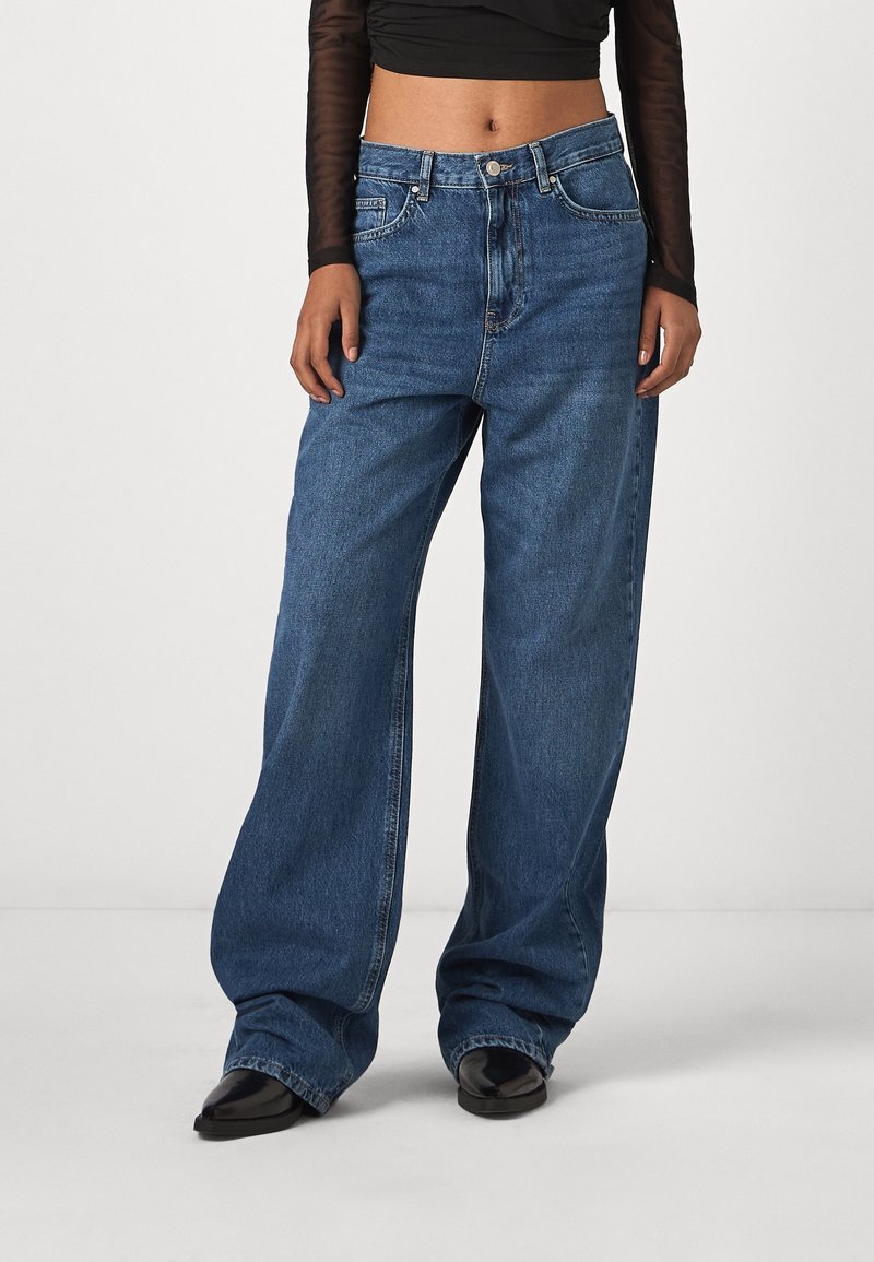 Personne portant un jean en denim bleu ample, des chaussures noires à bout pointu, et un haut noir à manches longues raccourci, debout devant un fond uni.