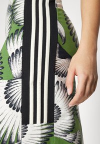 Vestido verde com um padrão de penas e riscas verticais em preto e branco; o material parece suave; mão repousando na peça.