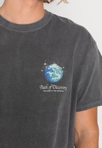 Grå bomullströja med en blå och grön jordgrafik med vita stjärnor, och text som lyder "Upptäcktsvägen, Universums kant."