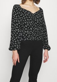 La blusa nera a fiori presenta un collo a cuore, maniche lunghe a sbuffo e un dettaglio arricciato sul davanti con piccoli motivi floreali bianchi e blu.