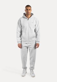 Muž stojící v světlém šedém zip-up mikině a odpovídajících sportovních kalhotách se bílými teniskami na jednobarevném světlém pozadí.