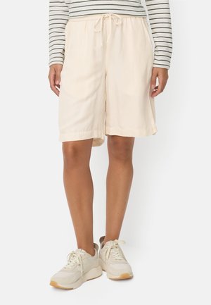 Person iført cremekrave, knælange løse shorts med snøre i taljen, stribet langærmet skjorte og lyse beige sneakers.