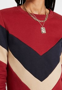 Personne portant un pull à motif chevron rouge, marine et beige, ainsi que des chaînes en or superposées avec un pendentif en forme de dragon rectangulaire.