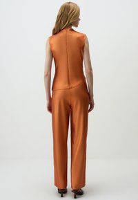 Satin oranje jumpsuit met mouwloze top, aangesloten pasvorm en wijde pijpen. Heeft een licht gebogen zoom en subtiele achternaaddetails.