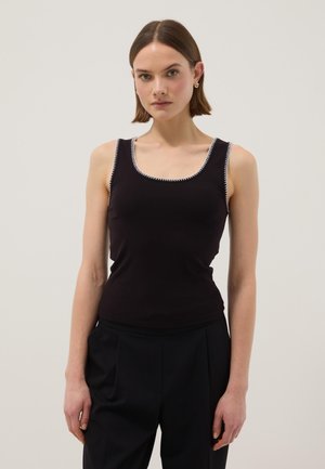 Junge Frau trägt ein schwarzes ärmelloses Top mit weißen Nähten an den Rändern und schwarze hoch taillierte Hose, steht vor einem einfarbigen Hintergrund.