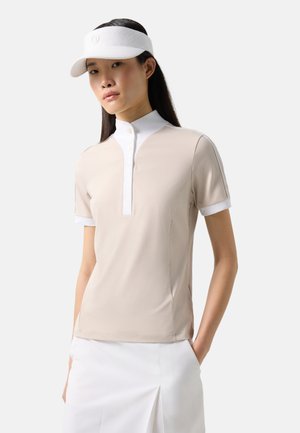 Vrouw draagt een beige poloshirt met korte mouwen, een witte kraag en manchetten, een witte rok met een voorsmouw, en een witte vizierpet, staand tegen een witte achtergrond.