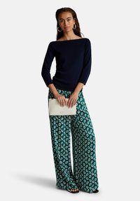 Top de manga larga azul marino combinado con pantalones amplios verdes de patrón con motivos de hojas blancas; sosteniendo un pequeño bolso claro.