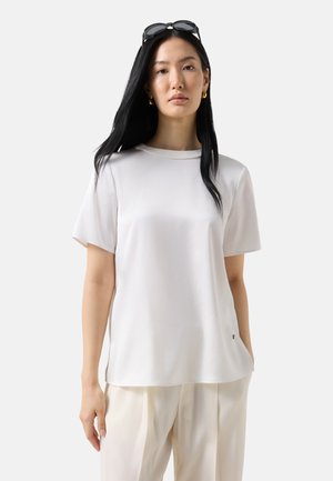 Woman with long black hair wearing sunglasses on her head, gold hoop earrings, white short-sleeve top, and cream pleated pants against white background.