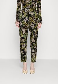 Pantalons noirs à motifs floraux avec des feuilles vertes et des fleurs blanches, fabriqués en tissu lisse, coupe fuselée, portés avec des chaussures plates de couleur claire.