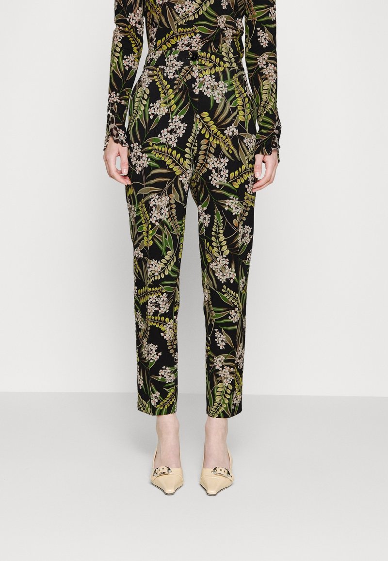 Pantalons noirs à motifs floraux avec des feuilles vertes et des fleurs blanches, fabriqués en tissu lisse, coupe fuselée, portés avec des chaussures plates de couleur claire.