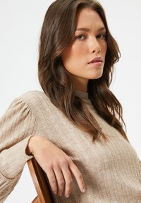 Beiger strukturierter Pullover mit Rippmuster, hohem Halsausschnitt und puffärmel. Das Model liegt mit der Hand auf einem Holzstuhl.