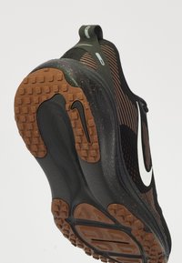 Nike athletic shoe featuring a black mesh upper, white swoosh, brown rubber outsole with textured grip, and a sleek, streamlined design.