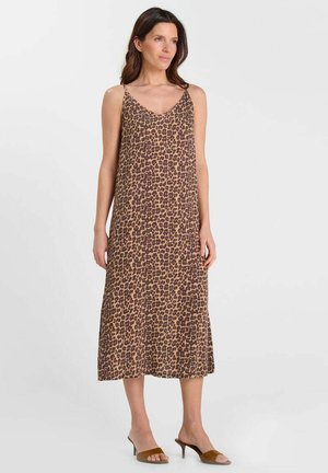 Vrouw die een mouwloze midi-jurk met bruine luipaardprint met dunne bandjes en bruine open-toe hakmuiltjes draagt, staand tegen een effen achtergrond.