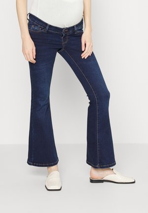 Osoba ubrana w ciemnoniebieskie, rozszerzane dżinsy, biały top i białe buty wsuwane, stojąca na tle jasnego, gładkiego tła.