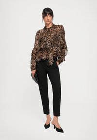Moteris vilkinti leopardų rašto permatomą marškinėlį, juodas siaurėjantis kelnes, juodas aukštakulnes basutes, laikanti juodą delninę, stovinti prieš baltą foną.