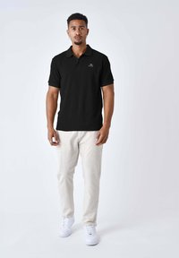 Camisa polo negra de canalé con mangas cortas, con un cuello y un pequeño logo. Combinada con pantalones de lino beige claros y zapatillas blancas.