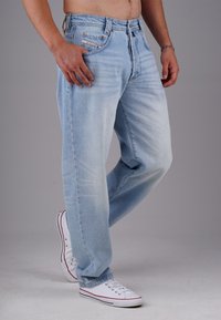 Hombre vistiendo jeans de denim azul claro y zapatillas blancas, de pie con una pierna ligeramente doblada contra un fondo gris liso.