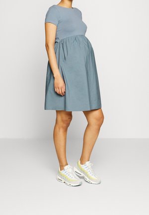 Zwangere persoon die een blauw jurkje met korte mouwen en witte sneakers draagt, staand tegen een effen lichte achtergrond.