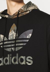 Svart huvtröja med en huva mönstrad i kamouflage. Har en stor, reflekterande Adidas-logotyp och tre ränder över bröstet.