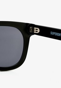 Zwarte zonnebril met grijze lenzen, vierkante montuur en een glanzende afwerking. Heeft zilverkleurige accenten bij het scharnier met "SUPERDRY" in blauw gedrukt.