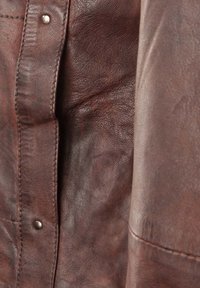 Bruine leren textuur met een zichtbare nerfpatroon, met een knoopdetail nabij de naad. Het oppervlak lijkt enigszins versleten en zacht.