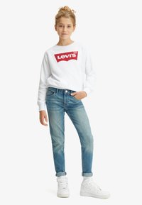 Hanorac alb cu logo roșu „Levi's ”, blugi bleu deschis și adidași albi. Croială casual, material moale din bumbac, manșete suflecate la blugi.