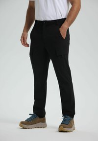 Mies, joka wear black cargo pants ja ruskeat vaelluskengät, joissa on siniset nauhat, seisoo toinen käsi taskussa yksinkertaista taustaa vasten.