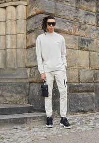 Béžová mikina s texturovaným vzorem, krémové cargo kalhoty s černými detaily a kapsami, černé tenisky, drží malou černou tašku.