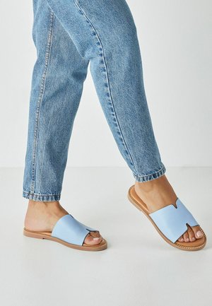 Light blue leather slide sandals feature a V-shaped cutout and a natural rubber sole, paired with light blue denim jeans.