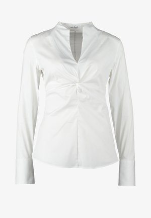 Wit shirt met lange mouwen en een gedraaide voorkant, gecombineerd met zwarte imitatieleren legging met geribbelde accenten. Zwarte clutch en hakken.