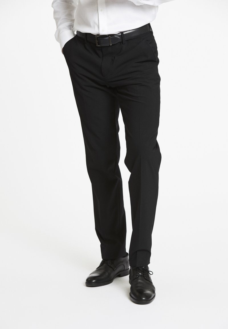 Zwarte op maat gemaakte broeken met een rechte pasvorm, subtiele textuur en voorplooien. Gecombineerd met zwarte leren dresschoenen en een wit shirt.