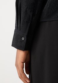 Gros plan d'une main à côté d'un poignet de chemise à manches longues noire avec de petits clous noirs et des détails de boutons noirs.