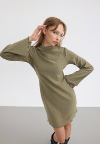 Olivegrünes langärmliges Kleid mit strukturiertem Muster und gewellten Bündchen, mit hohem Kragen und lockerer Passform. Schwarze kniehohe Stiefel.