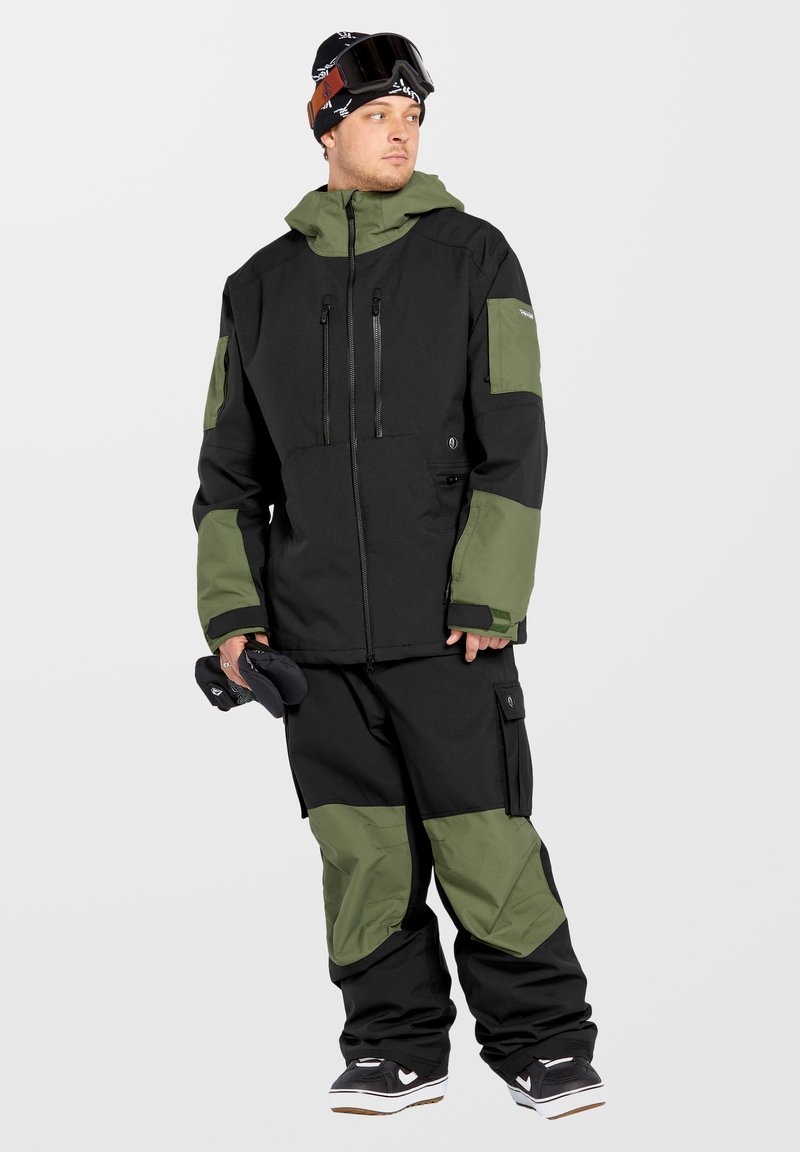Giacca da neve nera e verde oliva con cappuccio, tasche con zip e spalle rinforzate, abbinata a pantaloni da neve coordinati e scarpe nere.