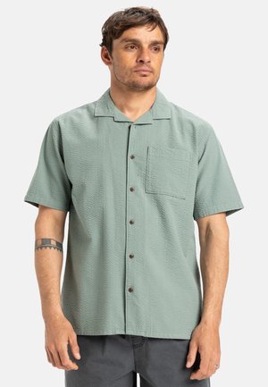Chemise à manches courtes boutonnée en tissu texturé vert clair, avec une poche poitrine et des boutons marron, idéale pour une tenue décontractée.