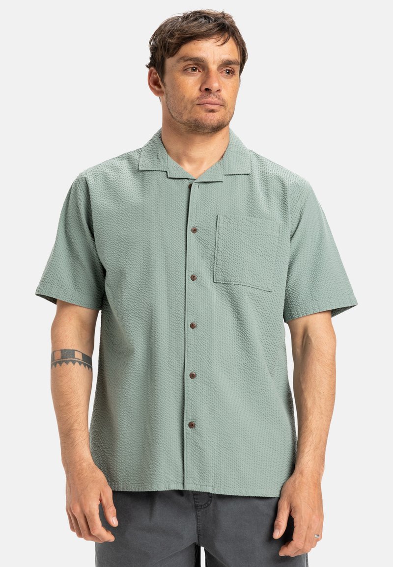 Short-sleeve button-up shirt in light green textured fabric, featuring a chest pocket and brown buttons, suitable for casual wear.