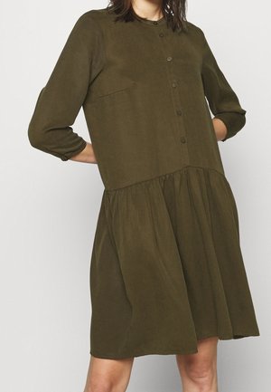 Frau trägt ein olivgrünes, langärmliges Hemdkleid mit geraffter Taille und rundem Ausschnitt.