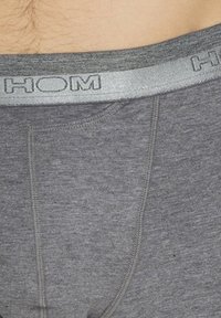 Undergarment grigio con una texture morbida, caratterizzato da un cinturino argento con il nome del marchio "HOM" in rilievo. Il design include dettagli di cucitura.