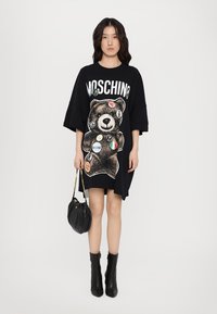 Μαύρο oversized φόρεμα μπλούζας με γραφικό αρκούδας, εκτυπωμένα μπαλώματα και κείμενο "MOSCHINO". Το μοντέλο κρατάει μια μαύρη τσάντα χειρός και φορά ankle boots.