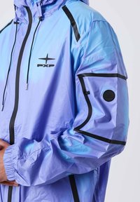 Persona che indossa una giacca a cappuccio lucida di un blu-viola chiaro con piping nero e logo PXP sul petto e sul braccio, con le mani leggermente serrate.