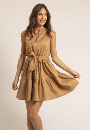 Femme souriante, vêtue d'une robe sans manches couleur beige avec un nœud ceinturé à la taille, tenant et faisant légèrement tournoyer la jupe sur un fond clair uni.