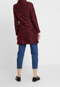 Cappotto a righe rosso e nero con colletto, maniche lunghe e tessuto testurizzato, abbinato a jeans blu con risvolto e scarpe nere.