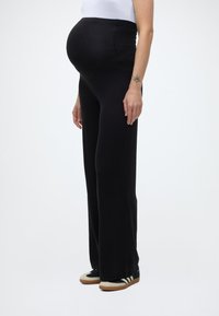 Raskaana oleva henkilö seisoo sivuttain yllään mustat äitiysfarkut, valkoinen paita, mustavalkoiset lenkkarit ja oikeassa käsivarressa rannetatuointi.