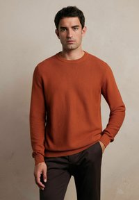 Musztardowy sweter z prążkowanej dzianiny, z okrągłym dekoltem i dopasowanymi mankietami, zestawiony z ciemnobrązowymi spodniami. Tkanina ma teksturowany wygląd.