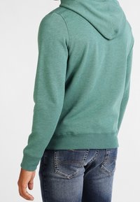 Sudadera verde con cordón, textura suave y puños acanalados. Combinada con jeans oscuros que cuentan con un diseño clásico de cinco bolsillos.