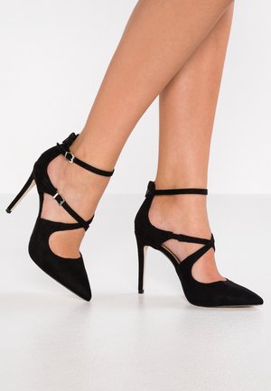 Czarne zamszowe szpilki z ostrymi noskami, z paskami wokół kostki i wyciętymi detalami po bokach. Posiadają smukły obcas typu stiletto.